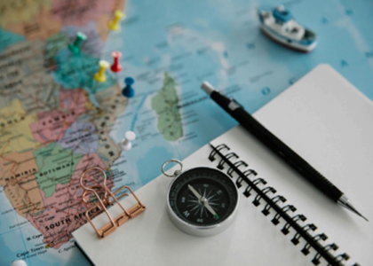 Picture of a brass compass pointing south, a notebook, and a black ballpoint pen, all placed on top of an unfolded map with several pins on different locations on the map.