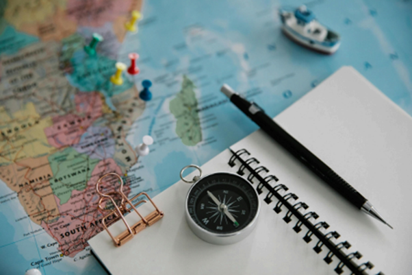 Picture of a brass compass pointing south, a notebook, and a black ballpoint pen, all placed on top of an unfolded map with several pins on different locations on the map.