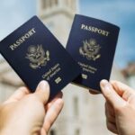 Two hands holding U.S. passports with a building in the background