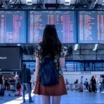 Picture of a lady wearing a flowery shirt and brown skirt with a slouchy denim book bag. She is looking at flight information on a big screen at a busy airport. Lots of other people involved in travel related activities are there too. The busy scene depicts the excitement of getting ready to travel.