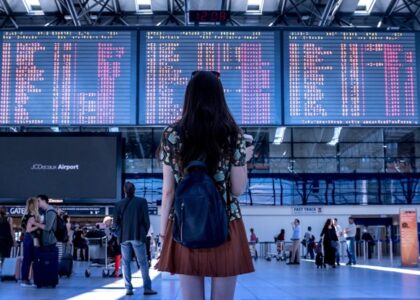 Picture of a lady wearing a flowery shirt and brown skirt with a slouchy denim book bag. She is looking at flight information on a big screen at a busy airport. Lots of other people involved in travel related activities are there too. The busy scene depicts the excitement of getting ready to travel.