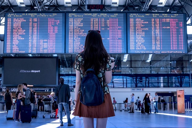 Picture of a lady wearing a flowery shirt and brown skirt with a slouchy denim book bag. She is looking at flight information on a big screen at a busy airport. Lots of other people involved in travel related activities are there too. The busy scene depicts the excitement of getting ready to travel.